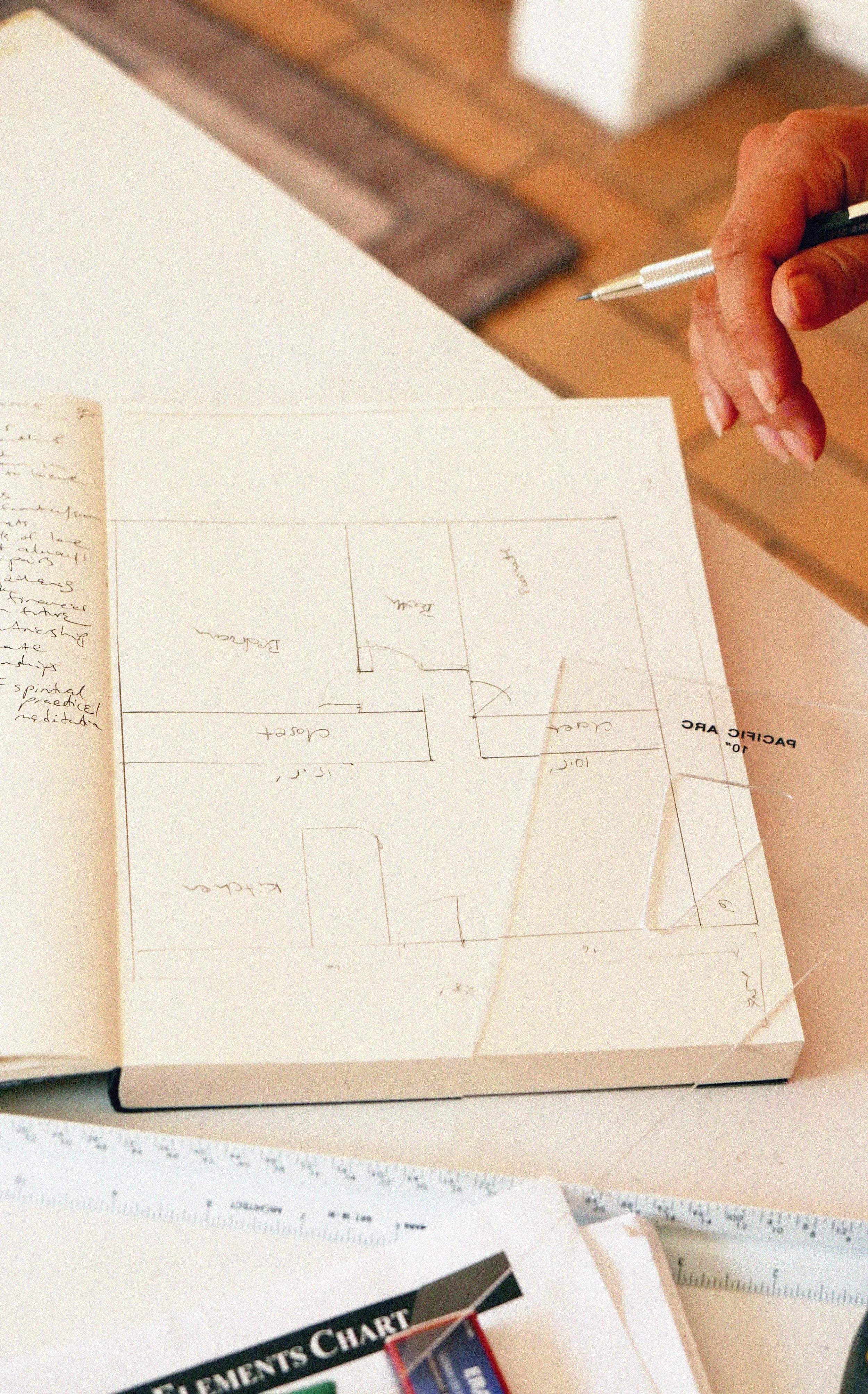 Hand holding a pen over a paper with a hand-drawn floor plan, notes, and measurements.