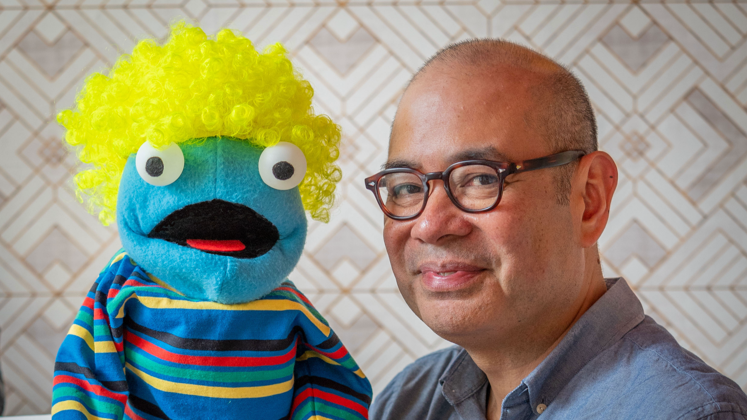 A man with glasses smiling next to a colorful puppet with yellow curly hair, big googly eyes, and a striped shirt, against a patterned background.