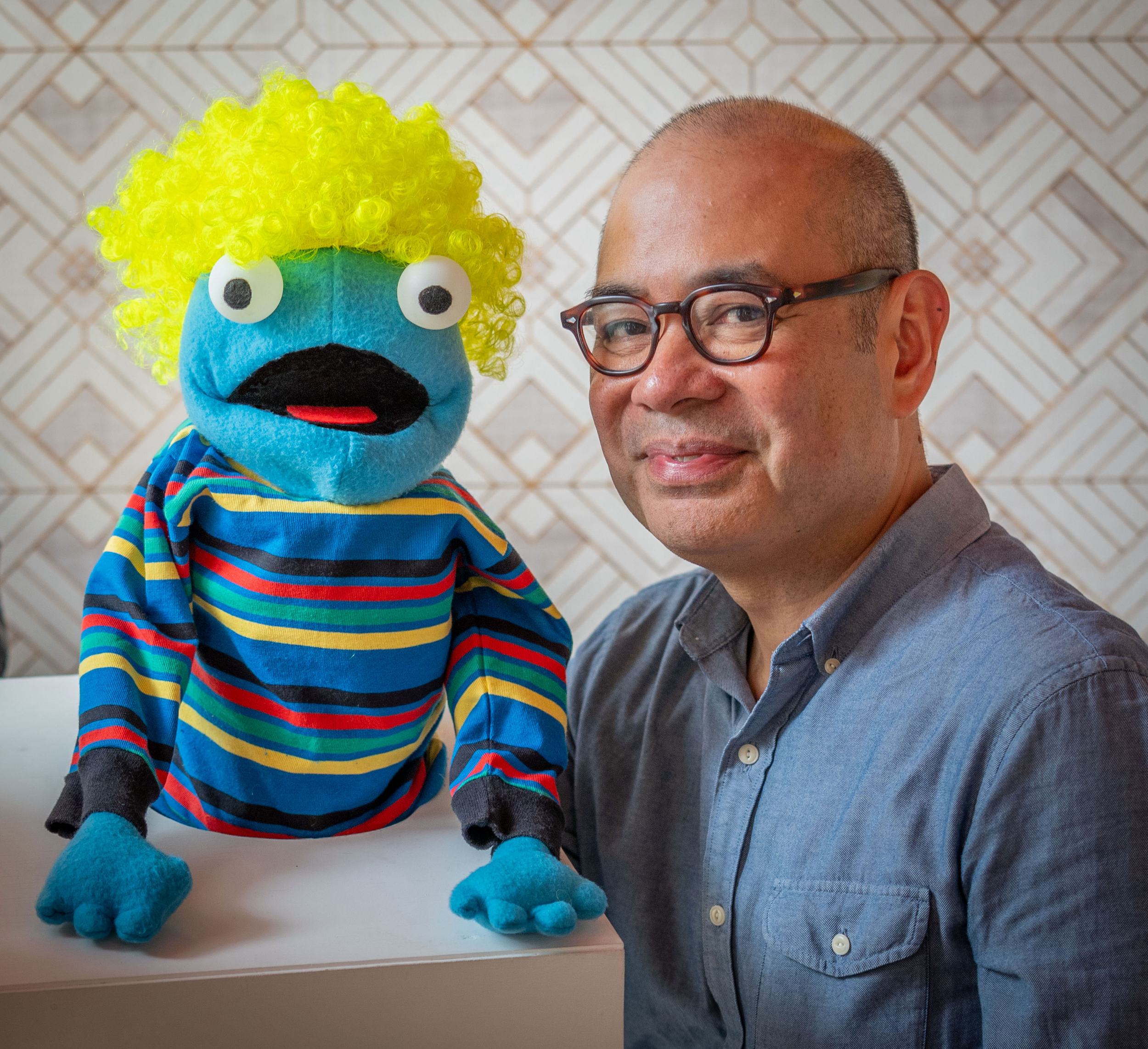 A man with glasses and a smiling expression is posing next to a colorful puppet with bright yellow curly hair, large googly eyes, a black mouth, wearing a striped multicolored shirt. They are indoors with a patterned wall in the background.