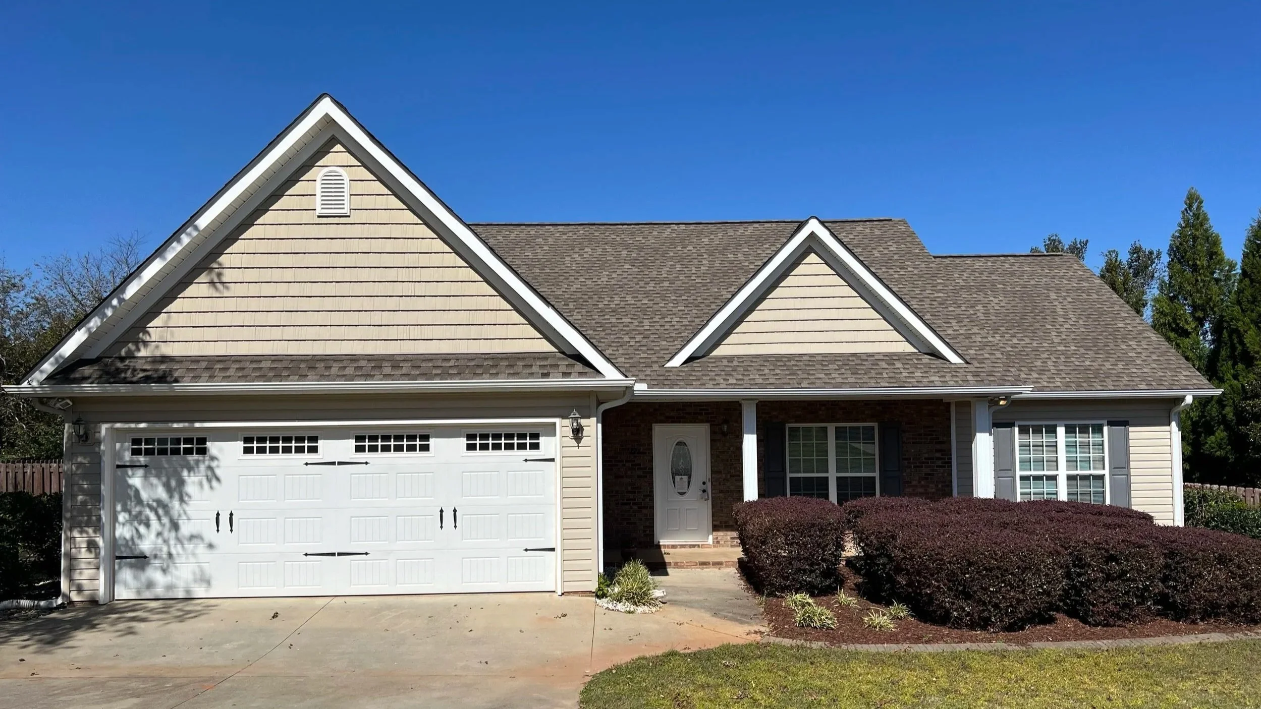 Front view of a modern suburban house with beige siding, a gabled roof, a white garage door, a white front door, and a landscaped yard with bushes and grass, under a clear blue sky.
