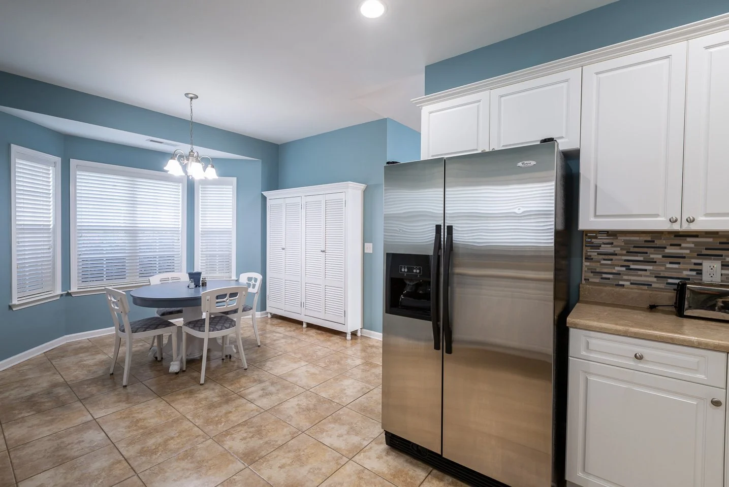 Kitchen with white cabinets, stainless steel refrigerator, beige countertops, tiled backsplash, tiled floor, white dining table with four chairs, windows with blinds, blue walls, and a chandelier.