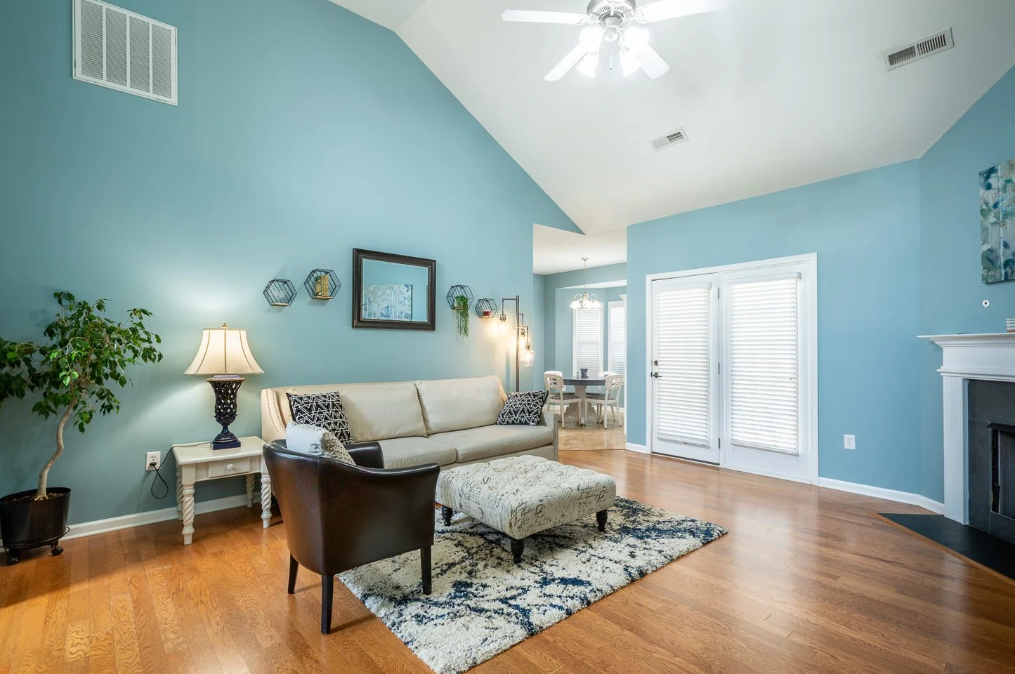 Living room with blue walls, white couch, black armchair, patterned ottoman, area rug, fireplace, and sliding glass doors with blinds.