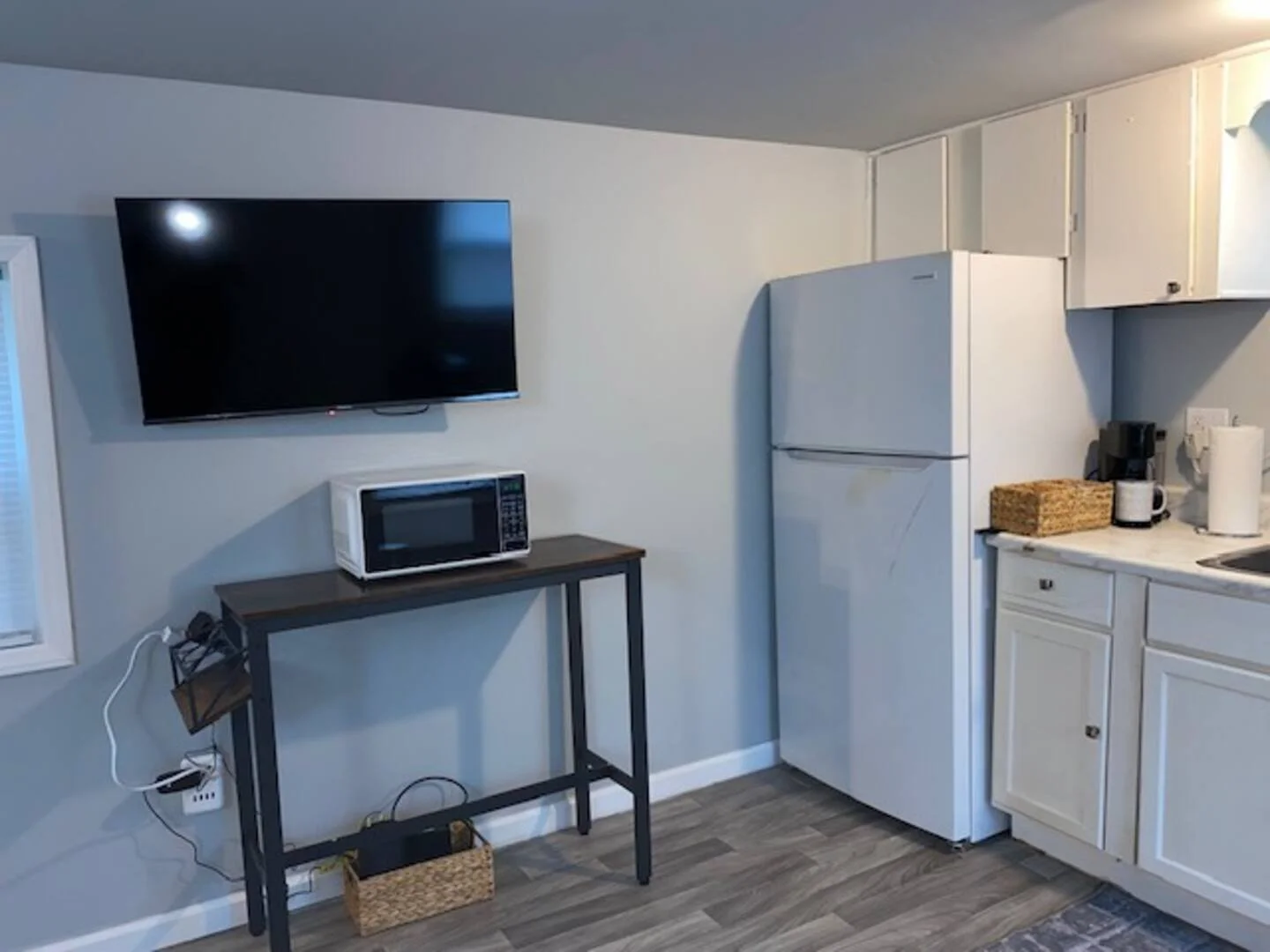 Kitchen area with a wall-mounted flat-screen TV, microwave on a black table, refrigerator, coffee maker, paper towels, and kitchen cabinets.
