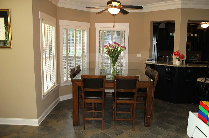 A dining room with a wooden table and six chairs, a vase with pink flowers, and three large windows with white blinds. A ceiling fan hangs above the table, and part of a kitchen with dark cabinets and a bar area with red flowers is visible in the bac