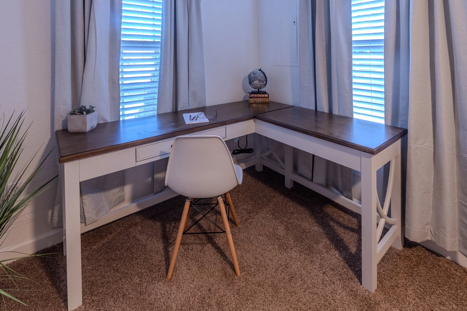 L-shaped desk with a dark wood top and white base, a white chair with wooden legs, window with blinds and curtains behind, small items on the desk including a pair of glasses, a globe, and a plant, brown carpeted floor.