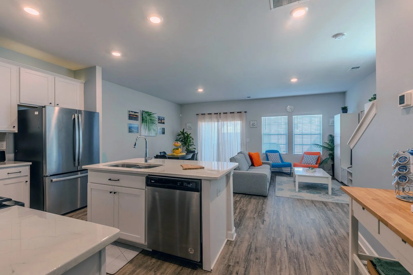 Open-concept kitchen and living room with modern decor, white cabinets, stainless steel appliances, gray sofa, colorful chairs, and natural light from large windows.