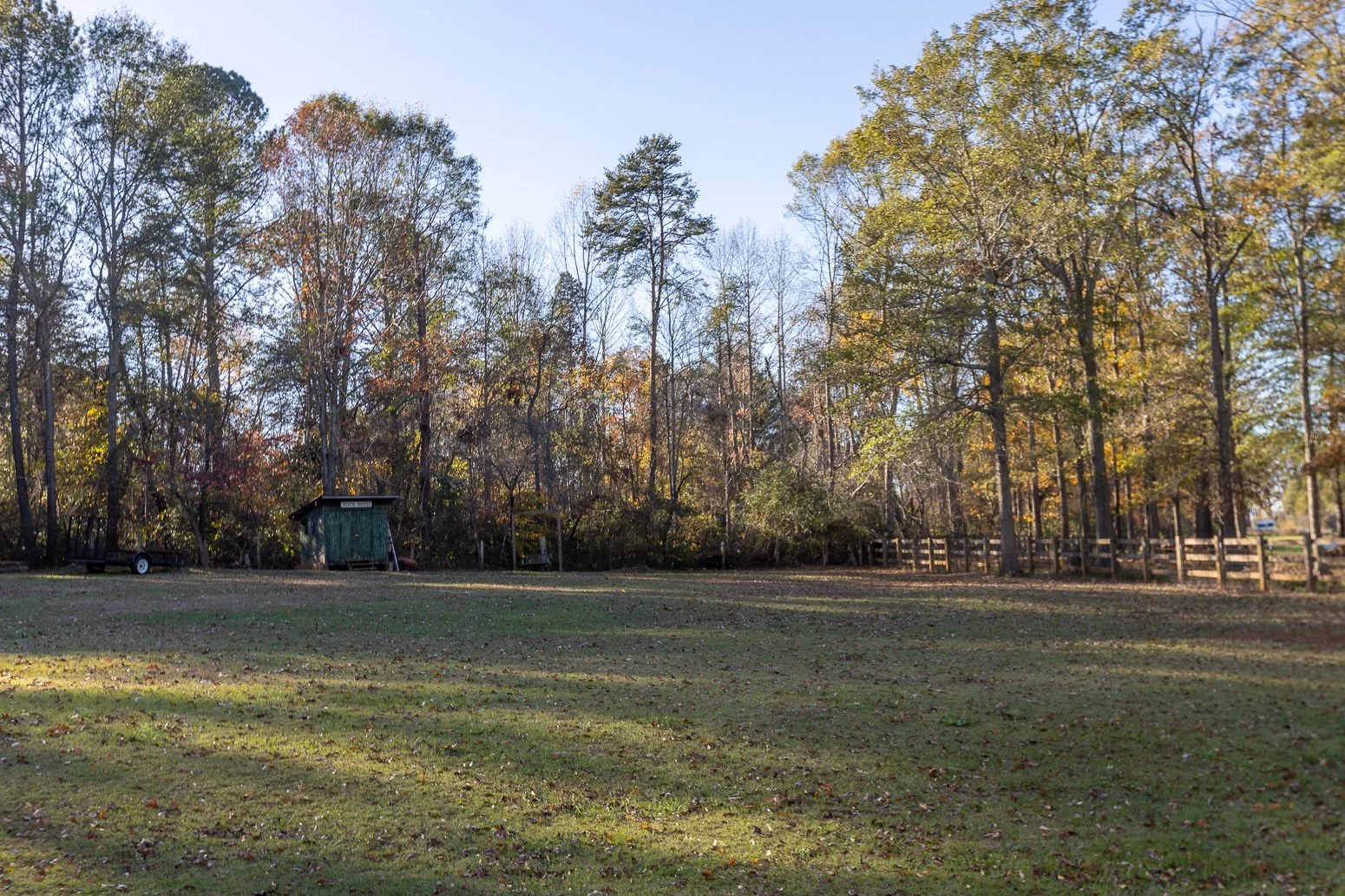 A grassy open field with fallen leaves, surrounded by trees with autumn foliage, a wooden fence on the right, and a small shed towards the left side of the background.
