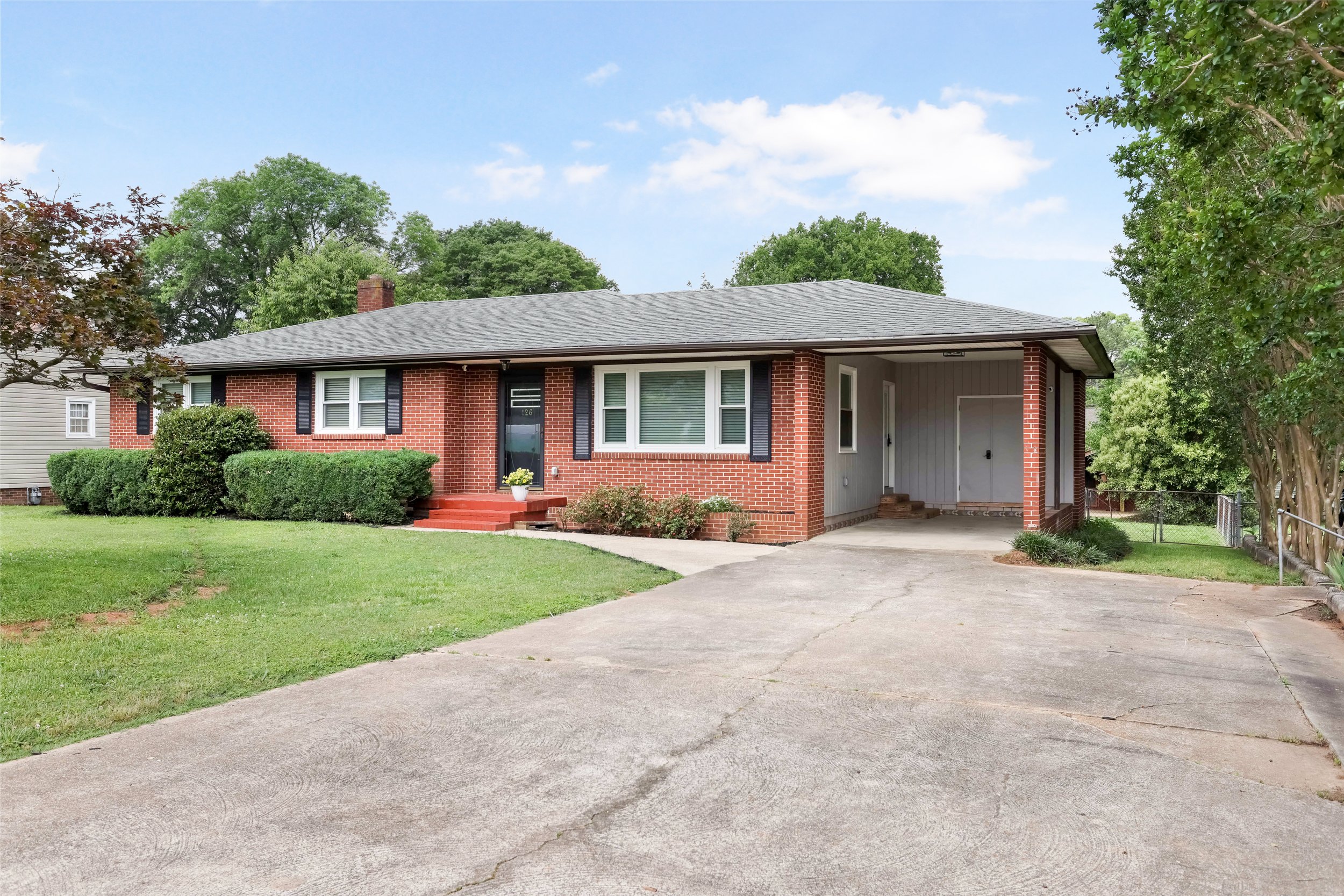 Front view of a single-story brick house with a gray roof, black shutters, a front porch with steps, green lawn, shrubs, and a driveway leading to a carport, surrounded by trees.