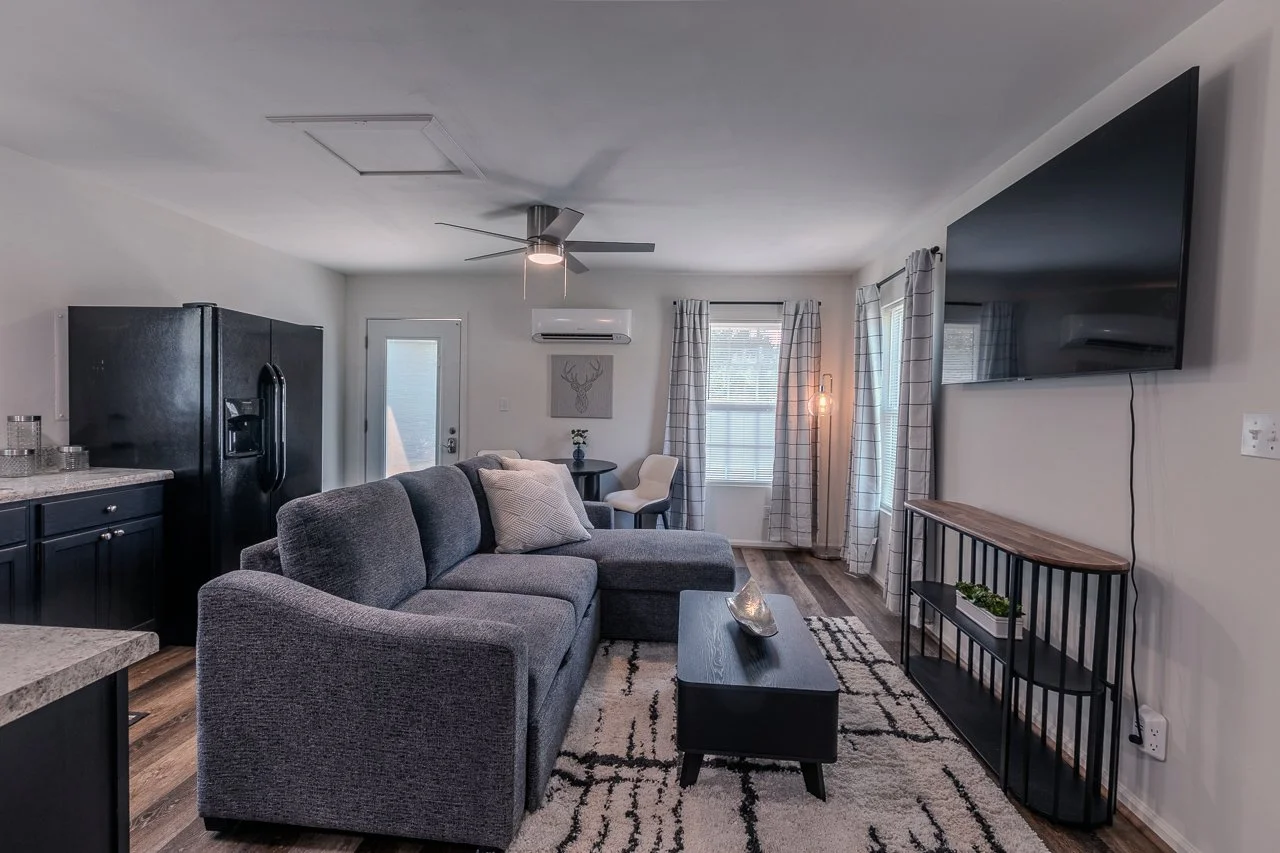 Living room with gray sectional sofa, black coffee table, patterned rug, wall-mounted TV, white curtains, and a small dining area with an air conditioning unit and wall art.