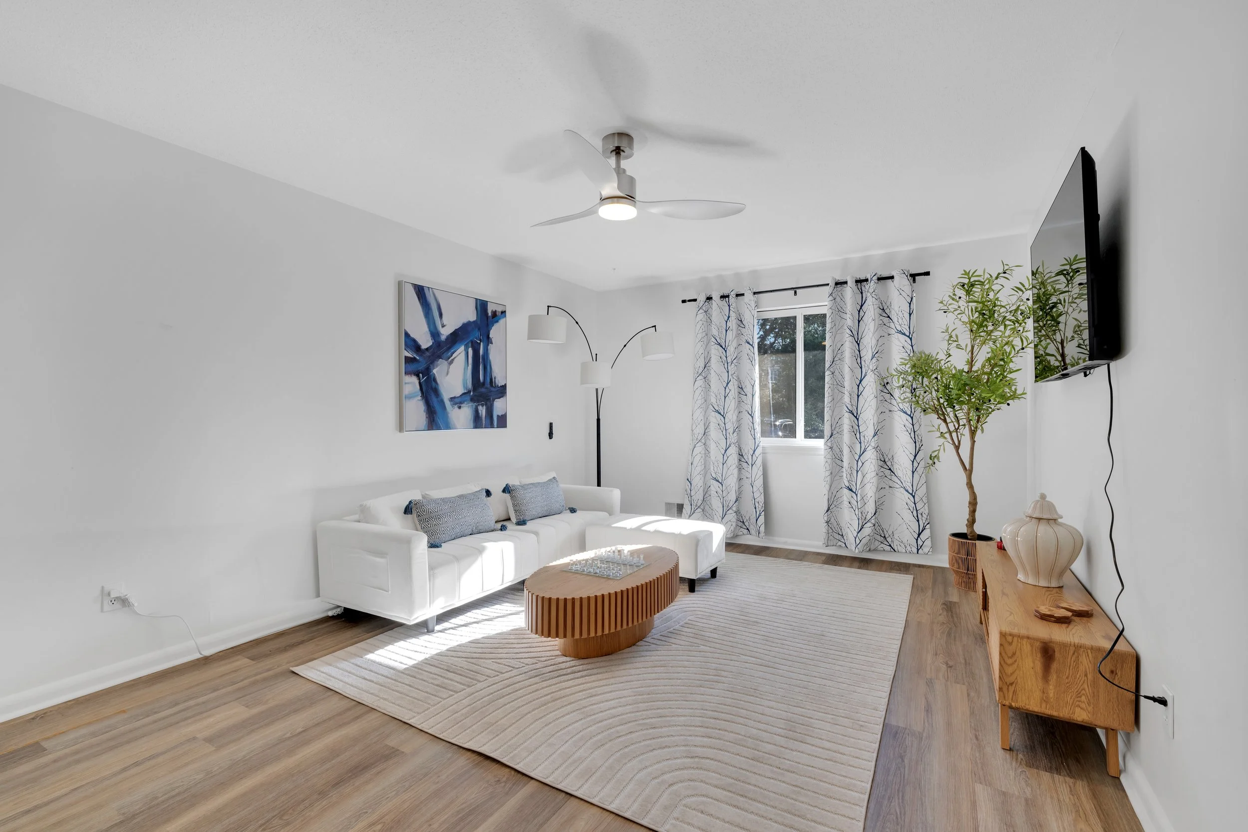 Living room with white walls, hardwood flooring, a white sofa with decorative pillows, a round wooden coffee table, a large abstract blue and white painting, a window with white and blue patterned curtains, a floor lamp, a tree in a pot, a wall-mount