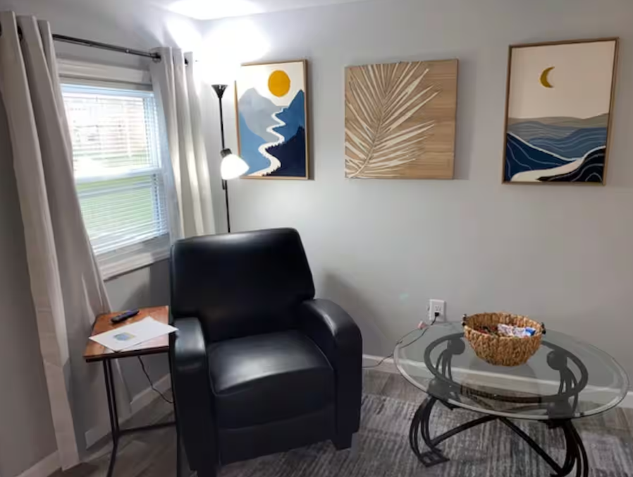 Living room with a black armchair, a round glass coffee table with a basket on it, a small side table with a remote and papers, a tall floor lamp, framed artwork on the wall, a window with white curtains, and a gray area rug.