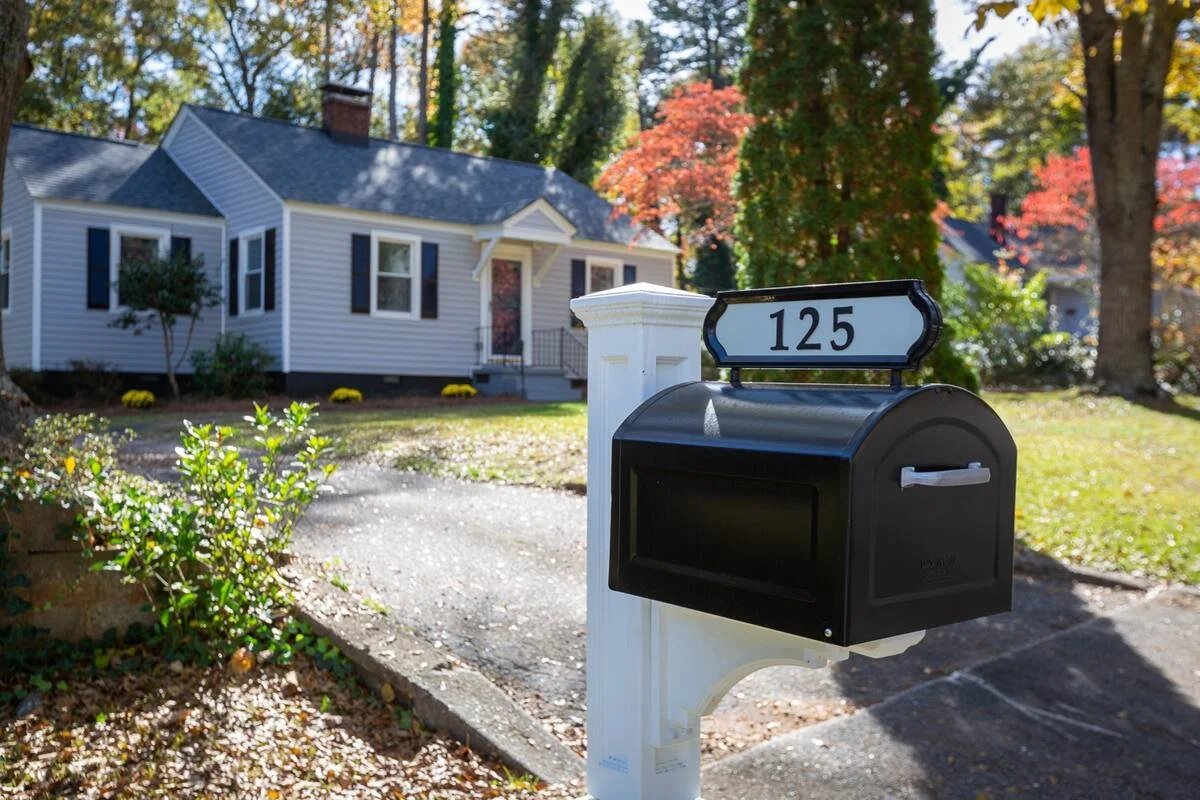 A black mailbox with a house number 125 on a white post in front of a white house with blue shutters and a front porch, surrounded by trees with fall foliage.