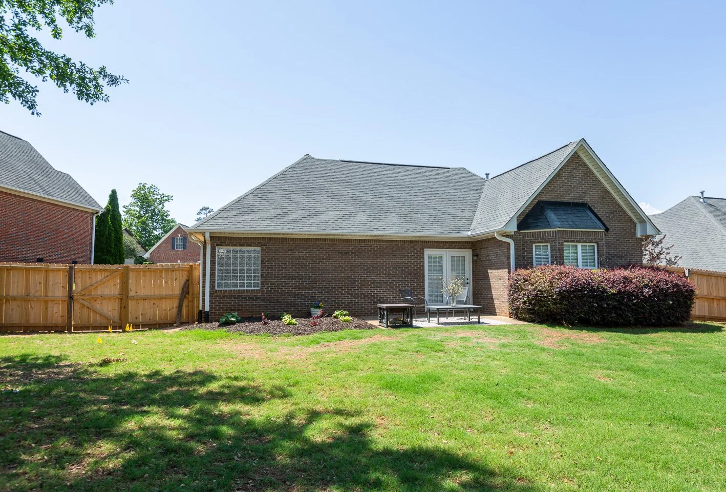 Backyard with green lawn, a small patio with furniture, and a brick house with French doors, surrounded by a wooden fence and some bushes under a clear blue sky.