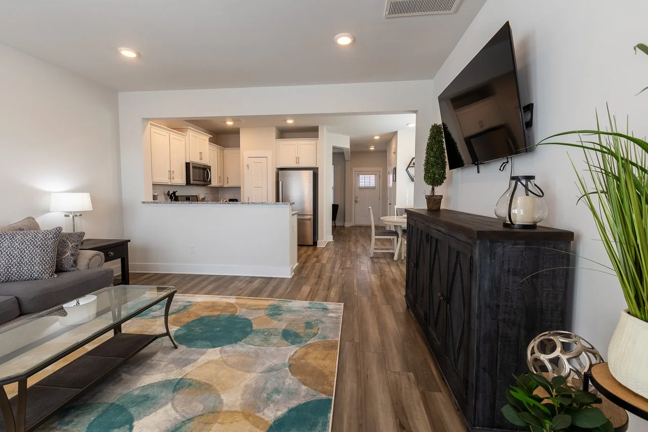 Living room with a gray sofa, glass coffee table, and a colorful rug. Flat-screen TV mounted on the wall above a dark wooden cabinet. There are plants and a lamp, with an open kitchen and dining area in the background.