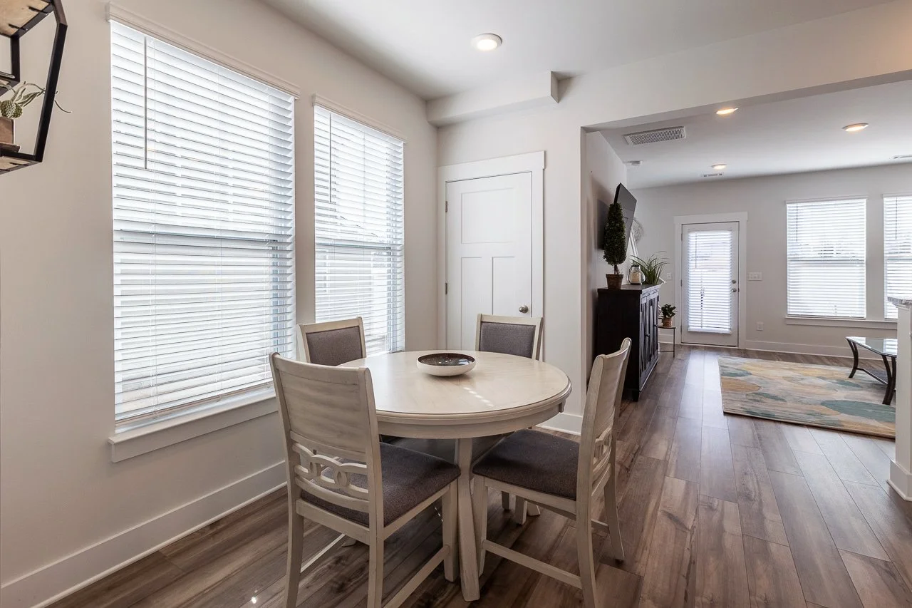 Dining area with a round table and four chairs, large windows with blinds, hardwood floors, and a decorative rug in an open-concept living space.