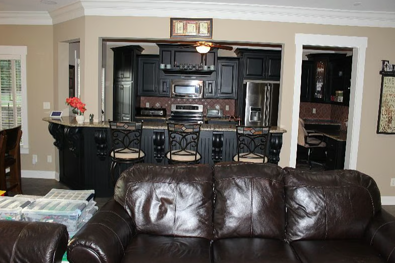 Living room with a dark brown leather sofa and a kitchen in the background featuring black cabinets, stainless steel appliances, and a breakfast bar with four barstools.