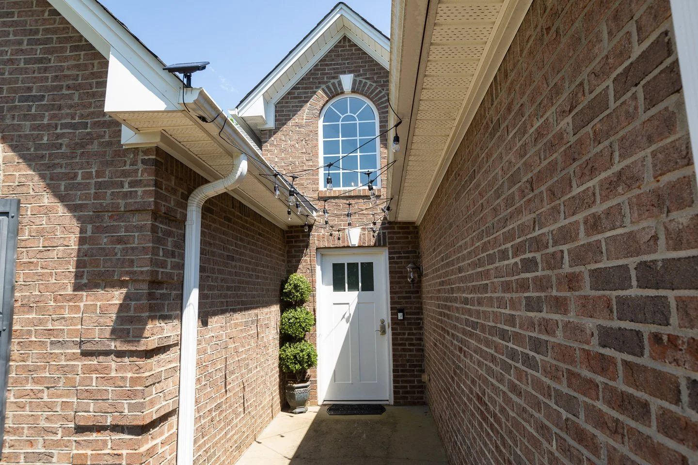 Narrow brick alley with a white door and decorative potted plant, string lights hanging overhead, exterior brick walls, and a brick house with a large arched window.
