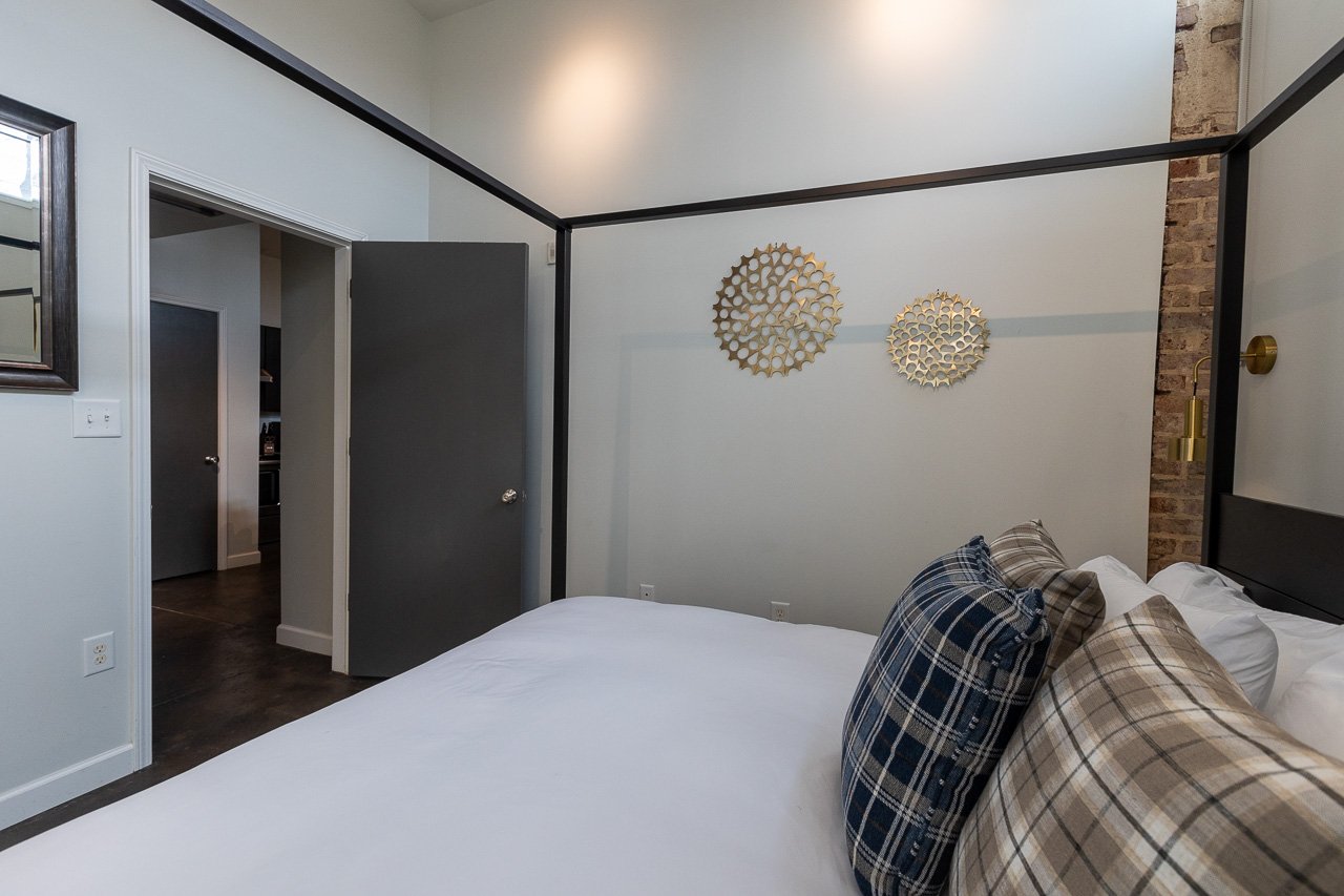 A bedroom with a white bed, pillows in plaid patterns, and wall decorations, with an open door leading to a hallway.