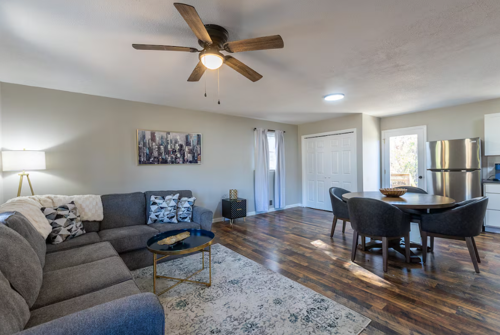 Living room with gray sectional sofa, throw pillows, a wood and gold coffee table, a floor lamp, wall art, hardwood flooring, a small black side table, a double-door closet, a curtained window, a dining table with four chairs, a stainless steel refri