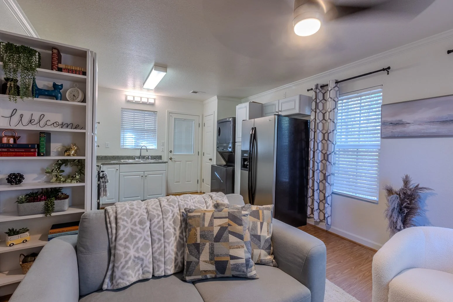 Living room with a beige couch, patterned pillows, a white bookshelf with decorative items, a kitchen area with white cabinets, a window, and a door in the background, wood flooring, and a window with patterned curtains.