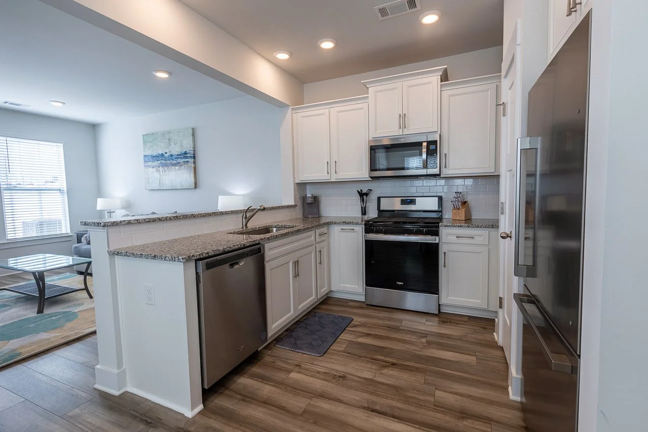 Modern kitchen with white cabinets, granite countertops, stainless steel appliances including a microwave, oven, dishwasher, and large refrigerator. There is a small gray rug on the wood floor and a view into a bright living room with a window, wall 
