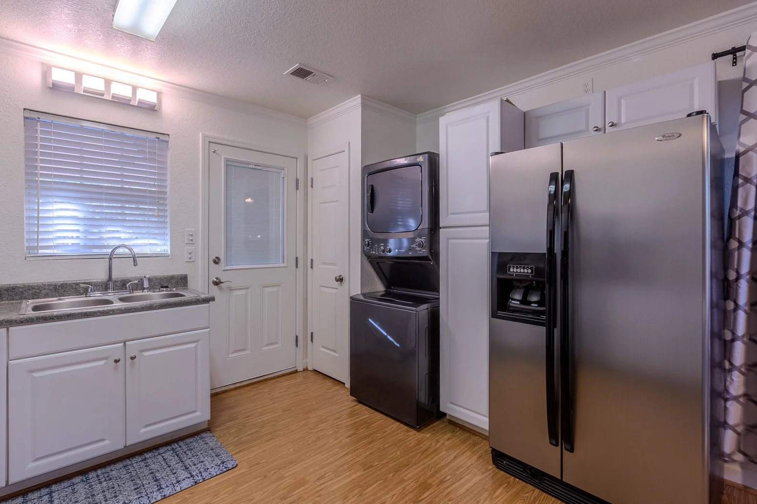 A laundry room with white cabinets, a double sink, a window with blinds, a door with a glass window, and a stainless steel refrigerator with a water dispenser, along with a stacked washer and dryer.