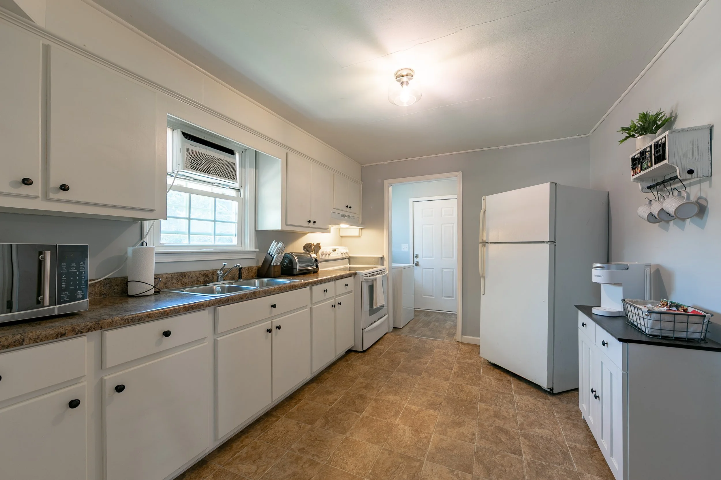 A kitchen with white cabinets, a window with an air conditioner, brown countertop, and appliances including a microwave, toaster, stove, refrigerator, and a washing machine. There is a small black counter with a white coffee maker and a wire basket. 