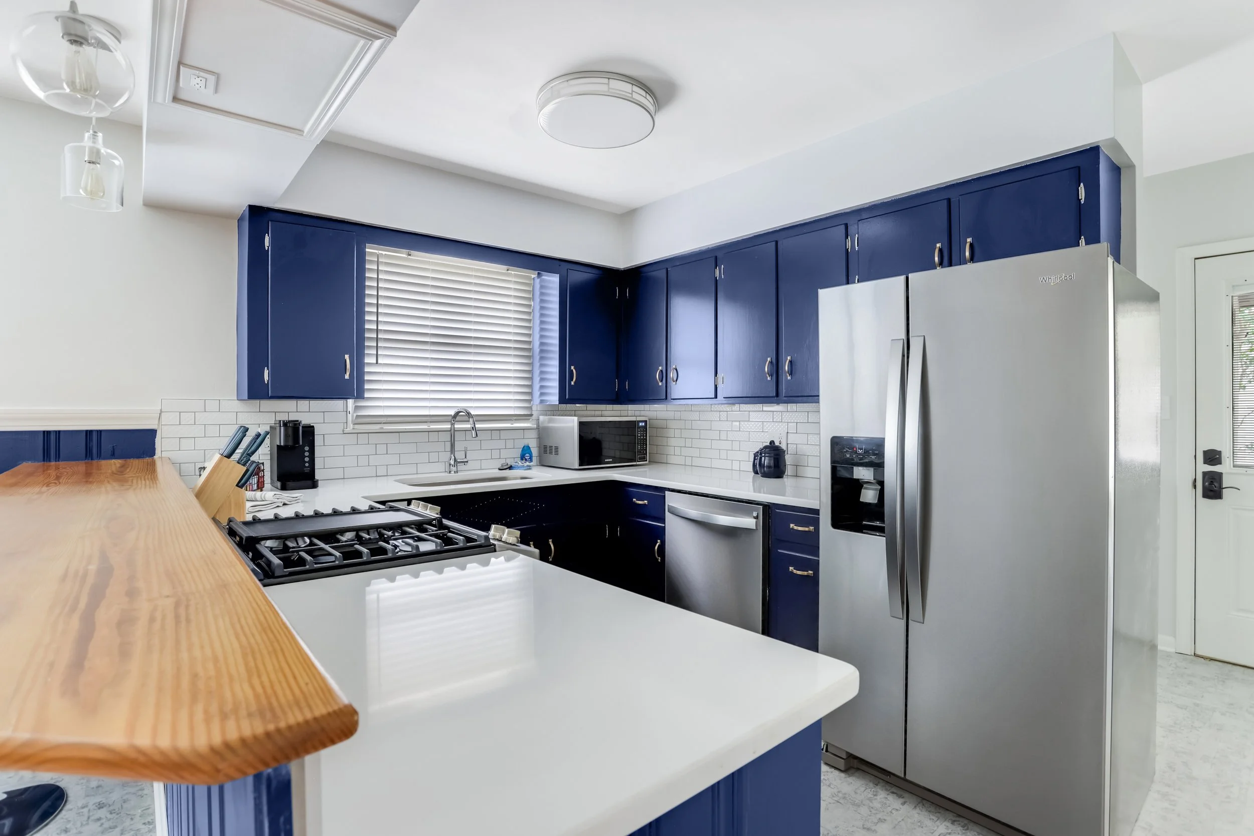 Kitchen with white countertops, blue cabinets, stainless steel refrigerator, microwave, and gas stove, with a window and white subway tile backsplash.