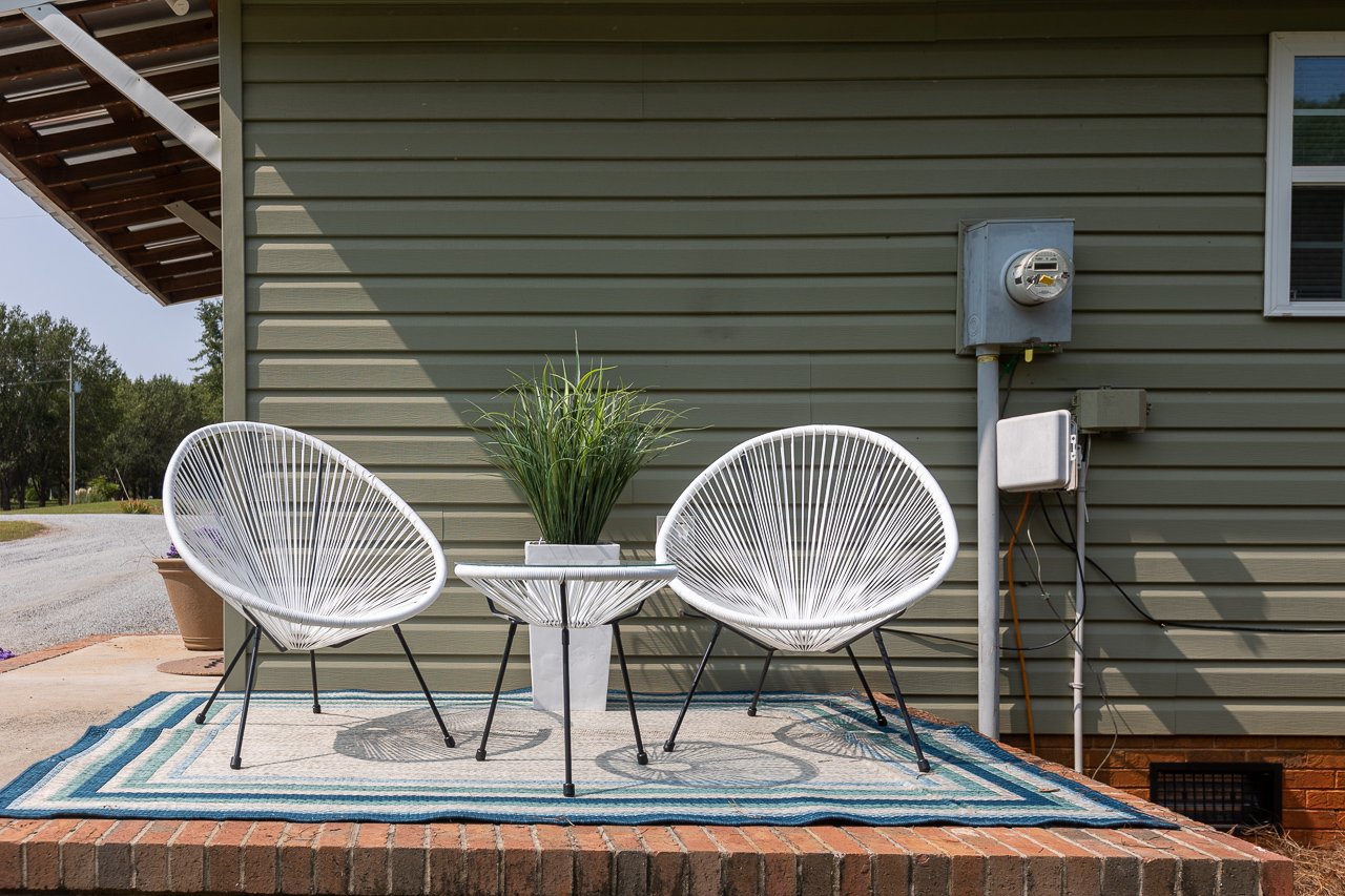 Outdoor patio with two white modern wire chairs, a small white table with green plants, a striped outdoor rug, against a gray house wall with electrical meters and conduit.