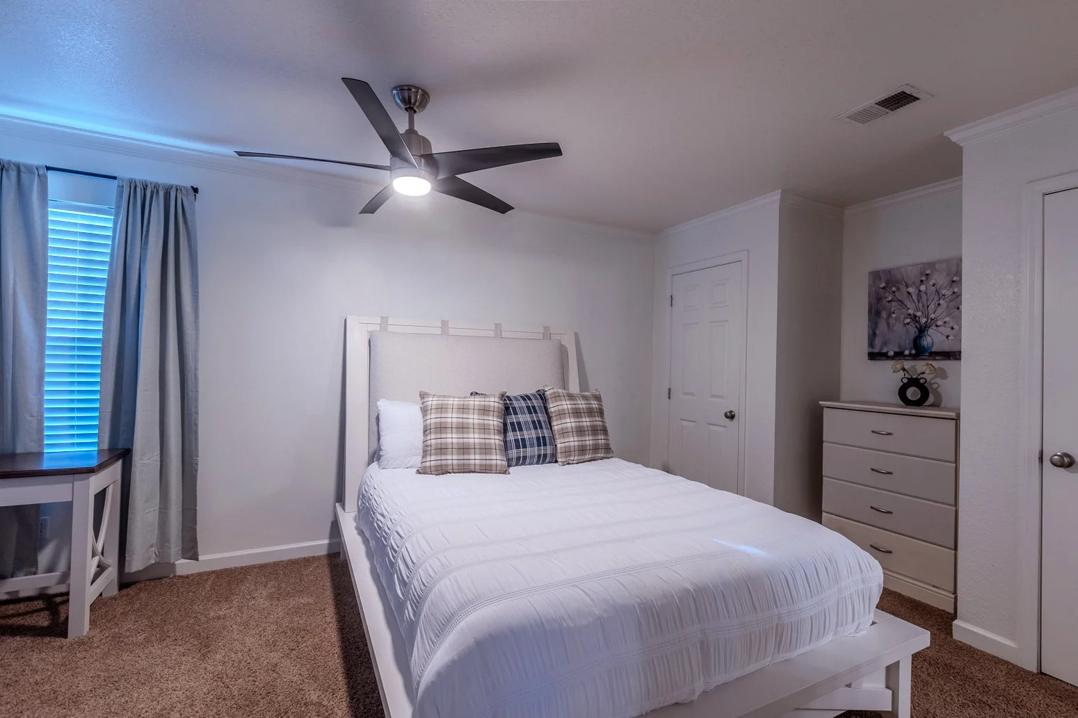 A bedroom with a white bed, pillows, a ceiling fan, a window with curtains, a dresser with a floral painting above it, and a door.