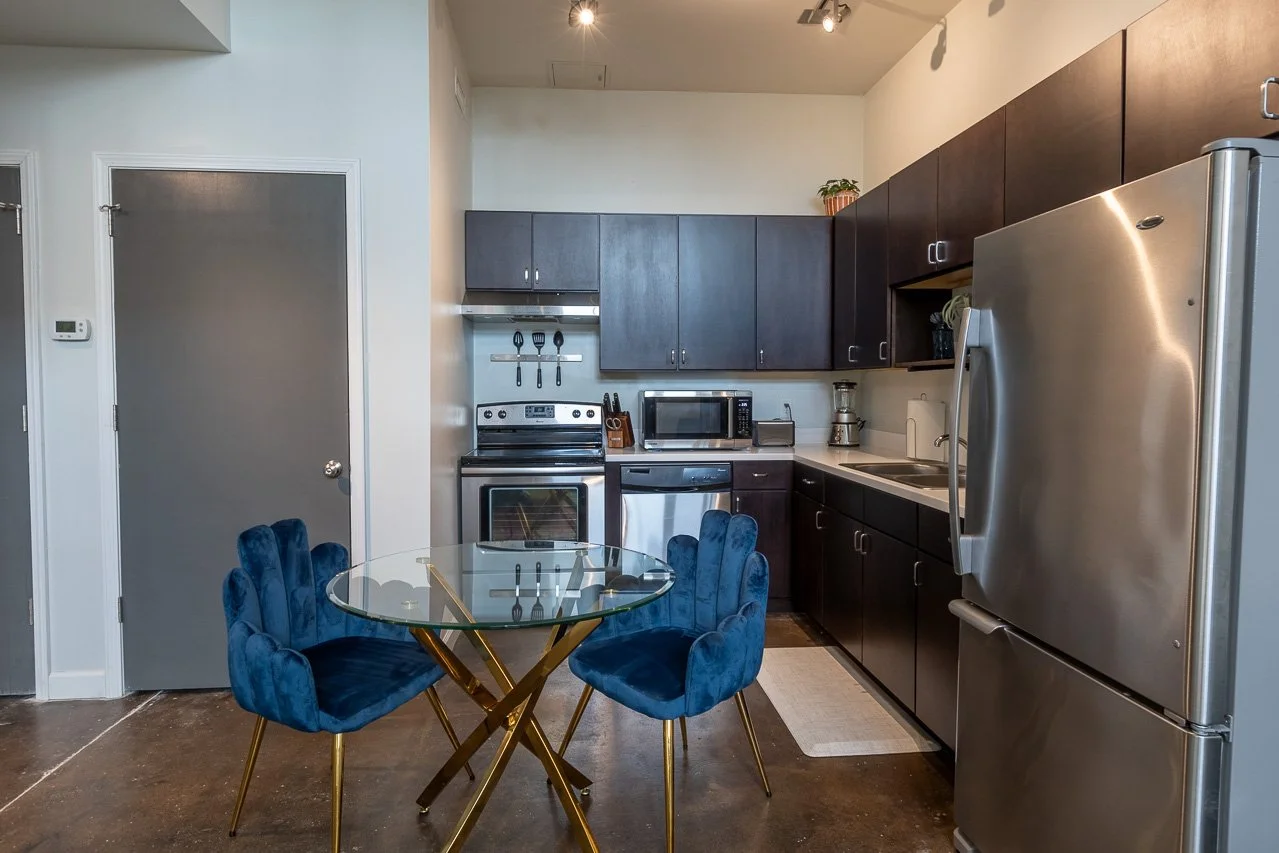 Modern kitchen with dark cabinets, stainless steel appliances including refrigerator, oven, microwave, and blender. Blue velvet chairs around a round glass table in the foreground.
