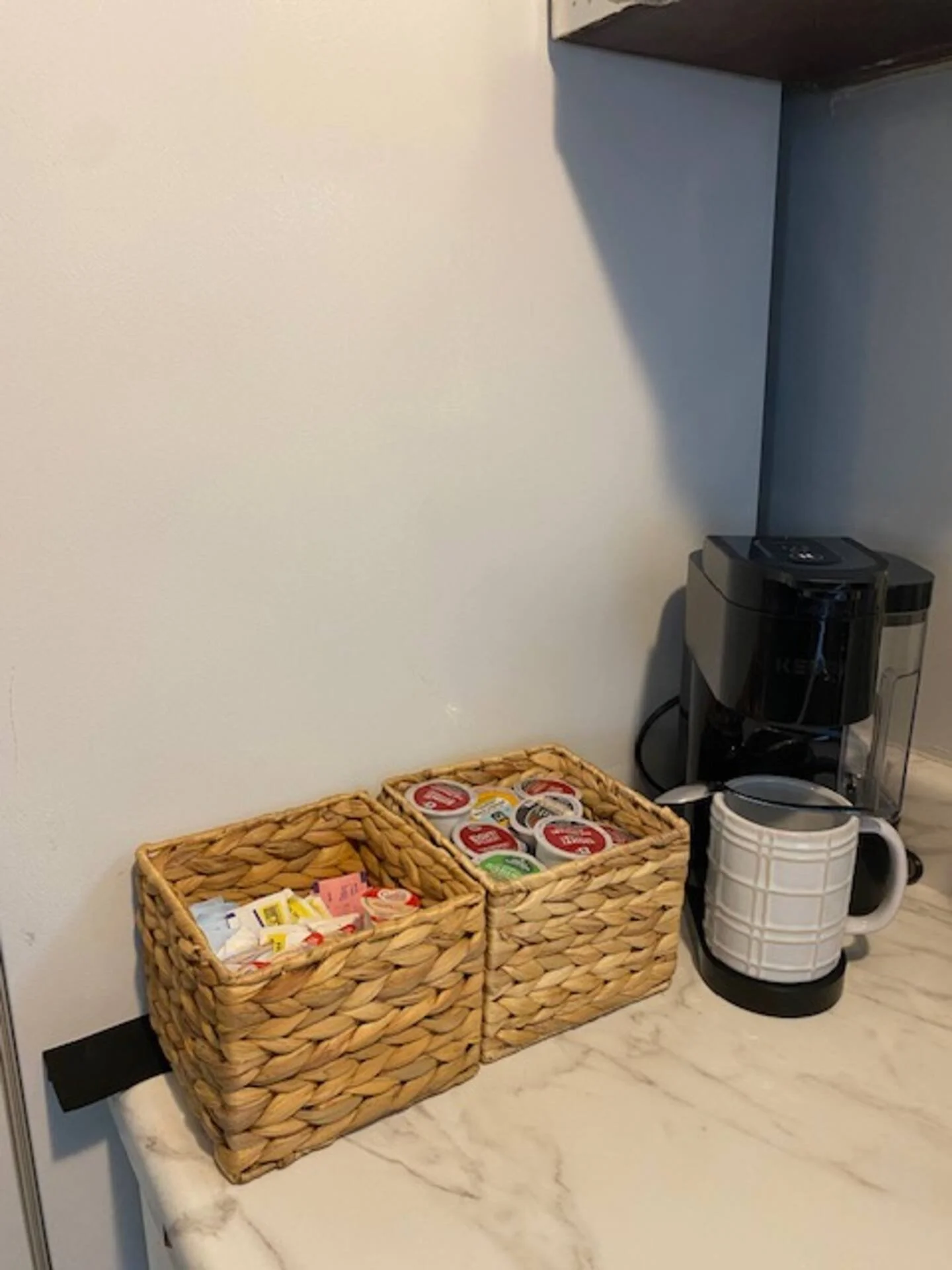 Coffee station with baskets of tea and coffee pods, a coffee machine, and a ceramic mug on a marble countertop.