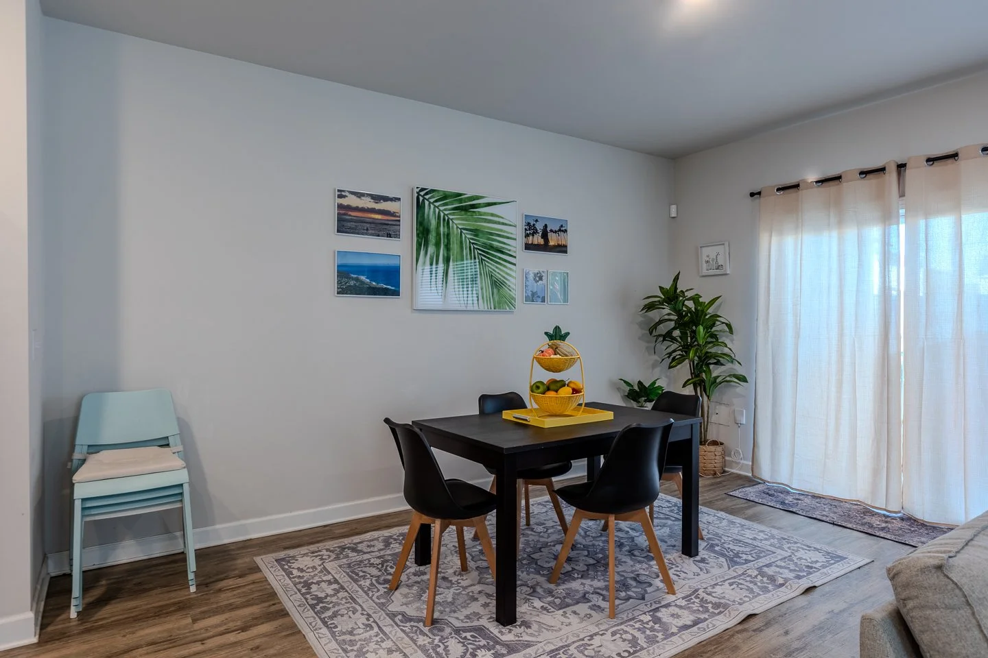Dining room with a black table and four black chairs, a yellow fruit basket centerpiece, wall decor with framed pictures and tropical art, potted plants near window with sheer curtains, light-colored rug on wooden floor.