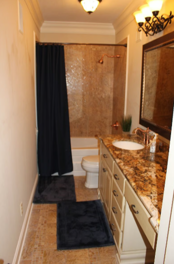 A small bathroom with a granite countertop vanity, a mirror, a sink, and a bathtub with a shower curtain, beige walls, and a ceiling light fixture.