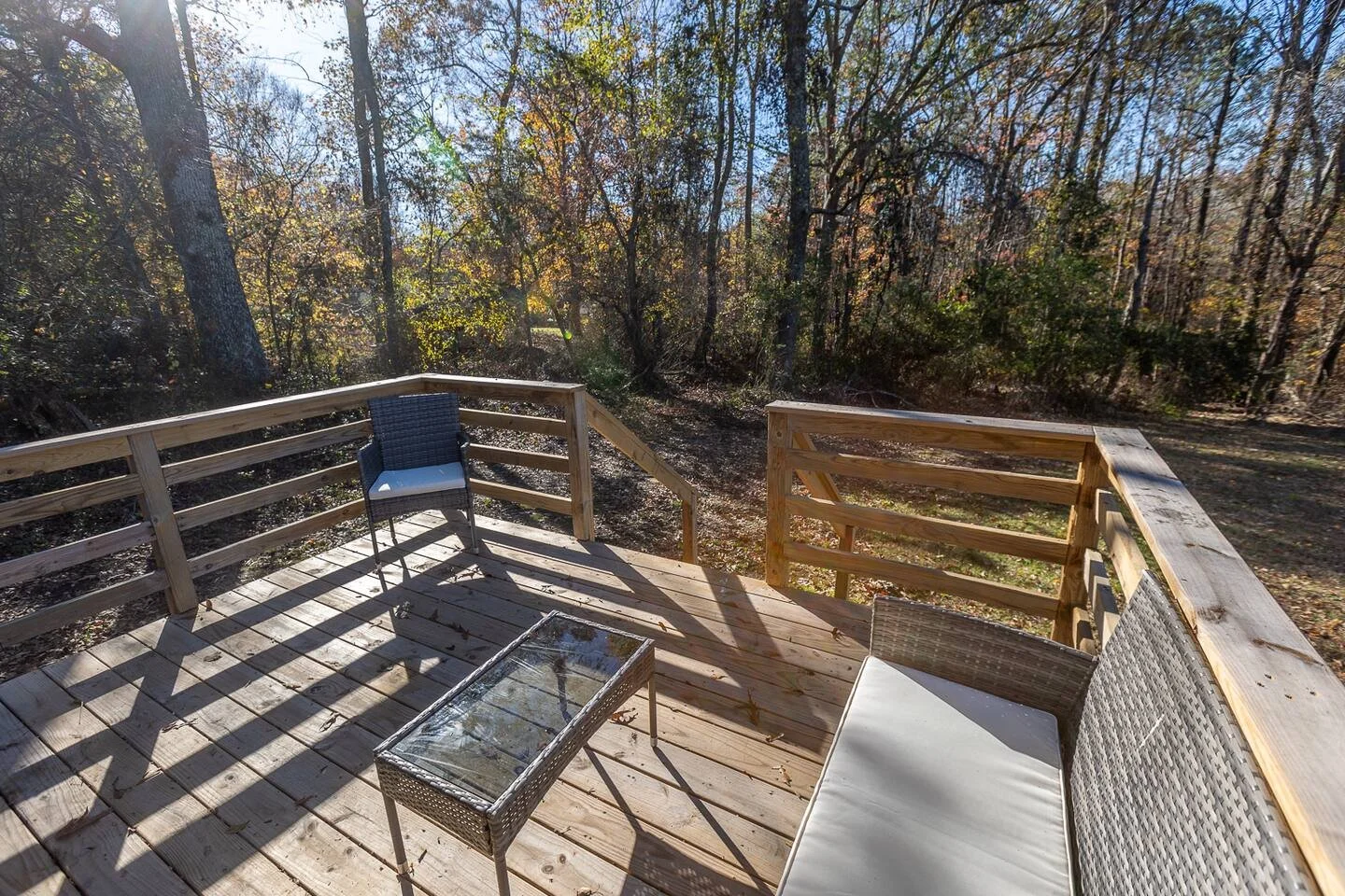 View of a wooden outdoor deck with outdoor furniture, including a glass-top table, a black chair, and a wicker loveseat with cushions. The deck overlooks a wooded area with trees and fall foliage.