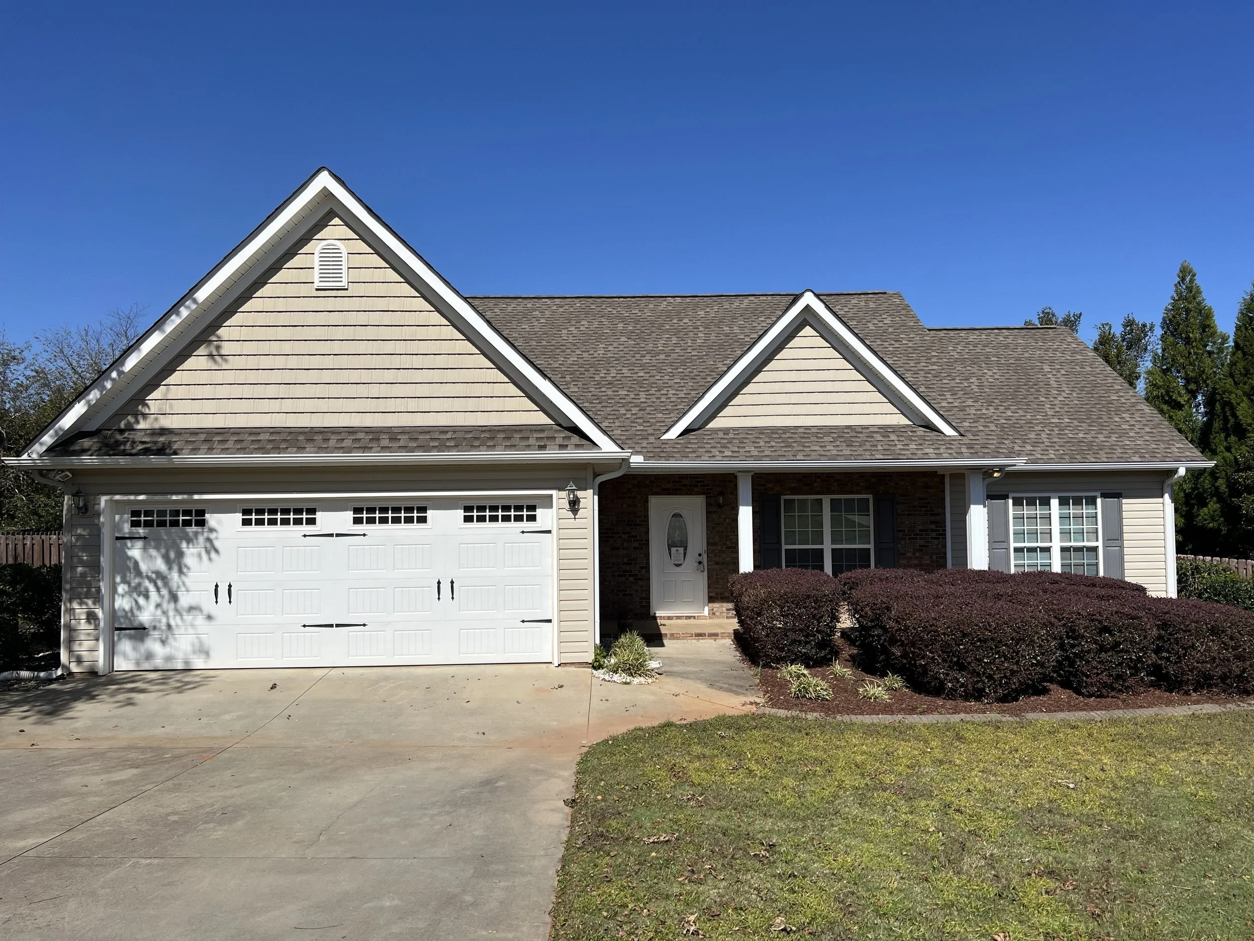 A suburban house with a two-car garage, beige siding, and brown roof, surrounded by bushes and a green lawn under a clear blue sky.
