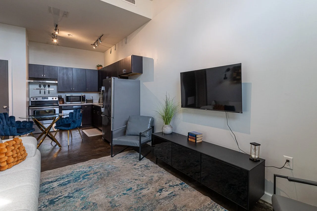 Modern living room with kitchen in the background, featuring a wall-mounted TV, gray armchair, black TV stand, decorative plant, and a glass dining table with blue velvet chairs.