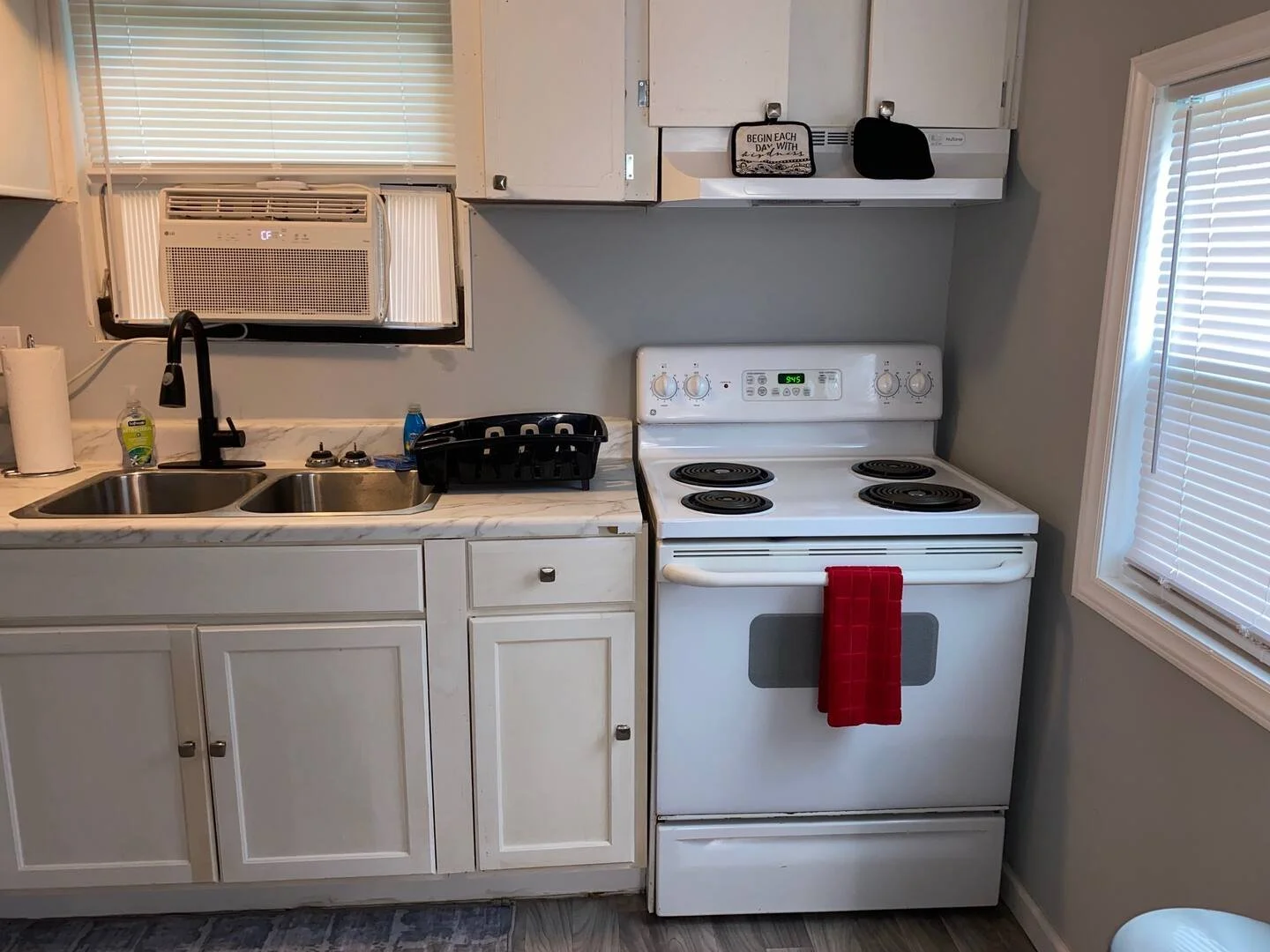 A kitchen with a double sink and a stove with four burners. There is an air conditioning unit in the window above the sink and a small window with blinds on the right. A red dish towel hangs on the oven handle, and there are two small black bags hang
