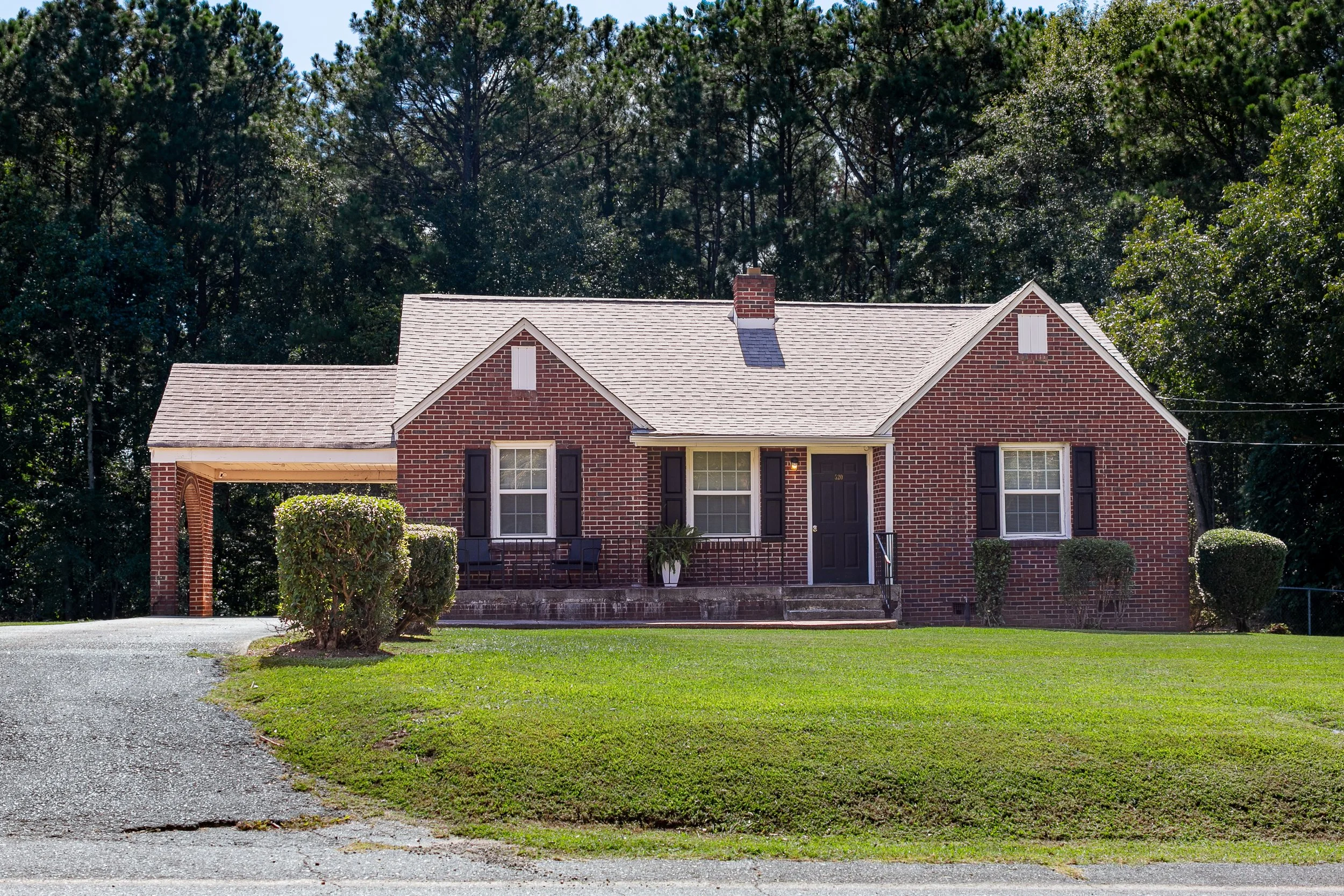 A brick house with a front porch, black shutters, and a side driveway, surrounded by a well-maintained lawn and trees in the background.