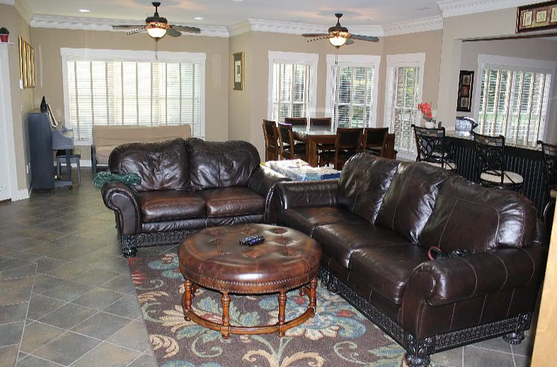 Living room with two dark brown leather sofas, a round leather ottoman with a remote on top, a patterned area rug, large windows with white blinds, a dining table with chairs, a piano, and a bar area with barstools. Ceiling fans are visible.