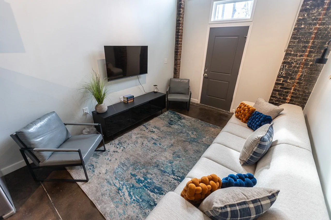 Living room with a white sofa, colorful pillows, a gray chair, a black TV stand with a flat-screen TV, a potted plant, and a textured rug on a dark hardwood floor, with exposed brick and a door in the background.