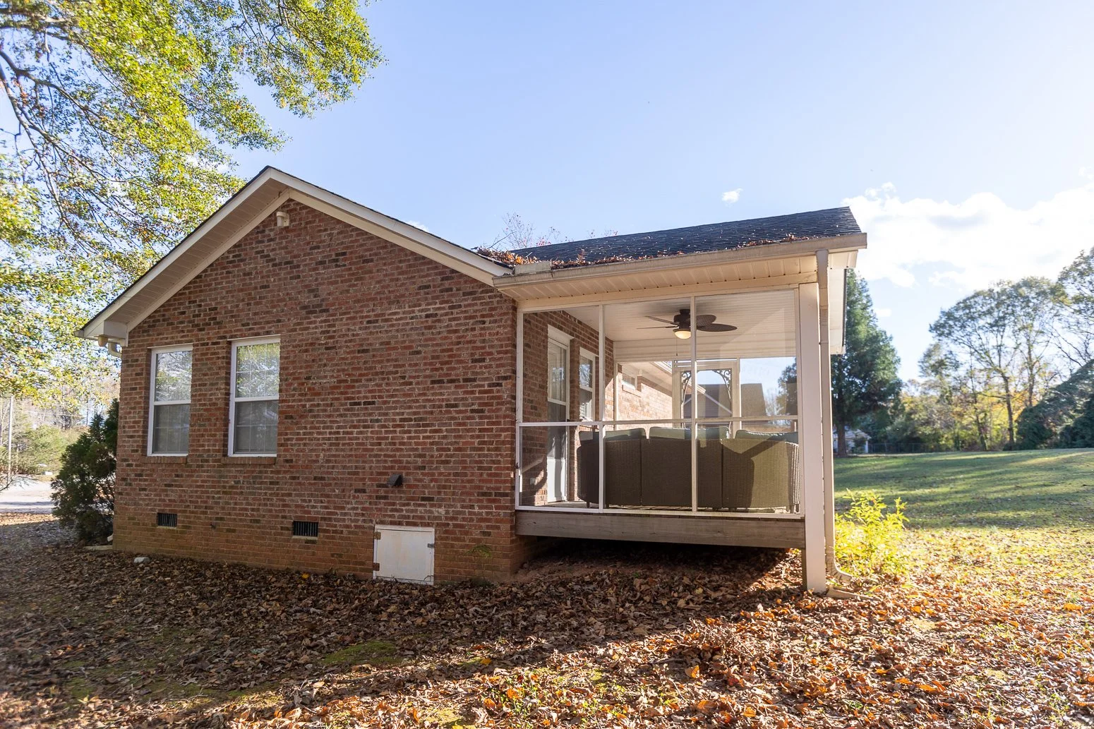 Back of a brick house with a screened-in porch and outdoor furniture, surrounded by trees and fallen leaves.