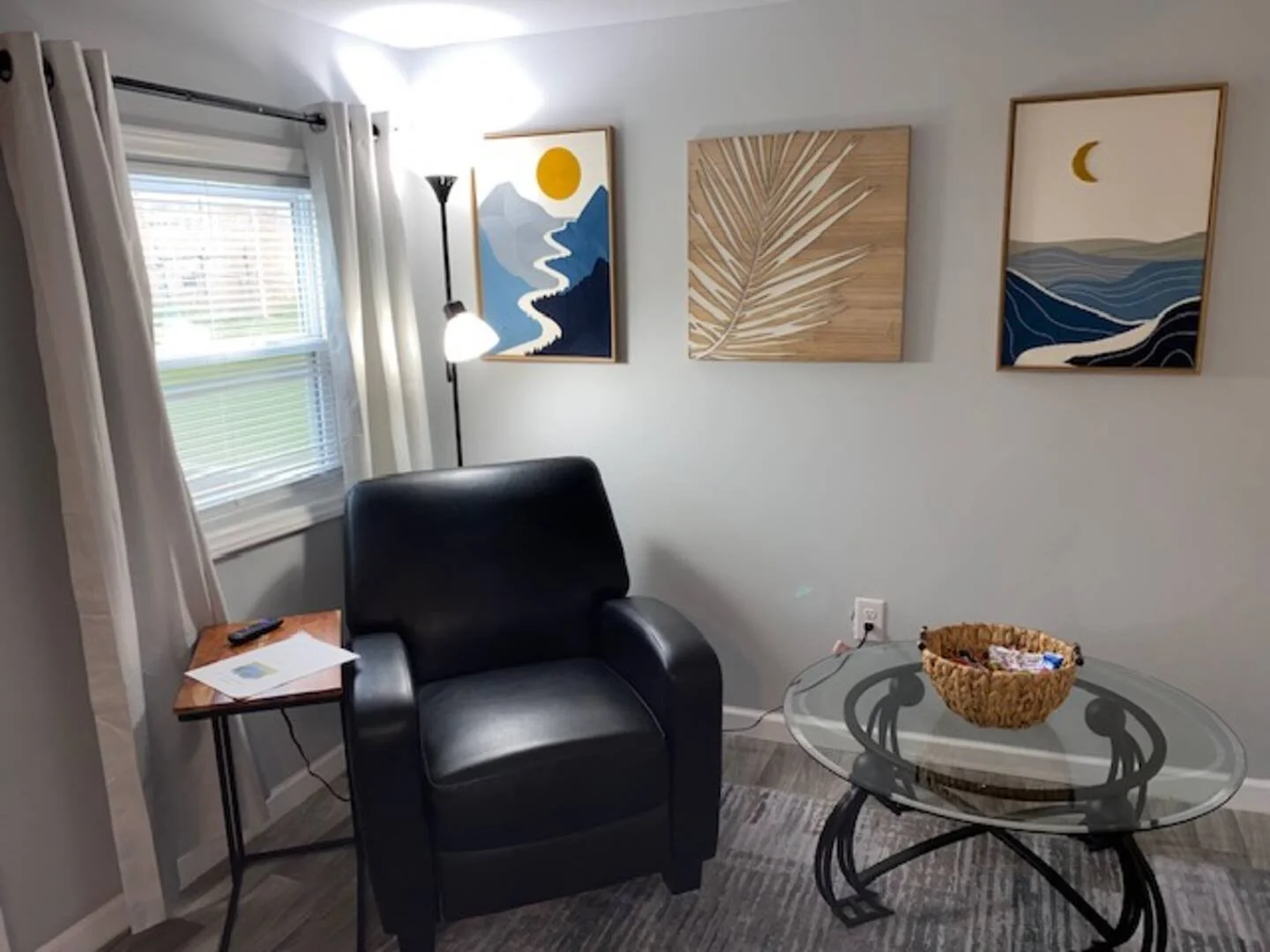 A cozy living room corner featuring a black leather armchair, a small wooden side table with a remote and papers, a glass coffee table with a woven basket, a window with white curtains, a standing lamp, and three abstract nature-themed paintings on t