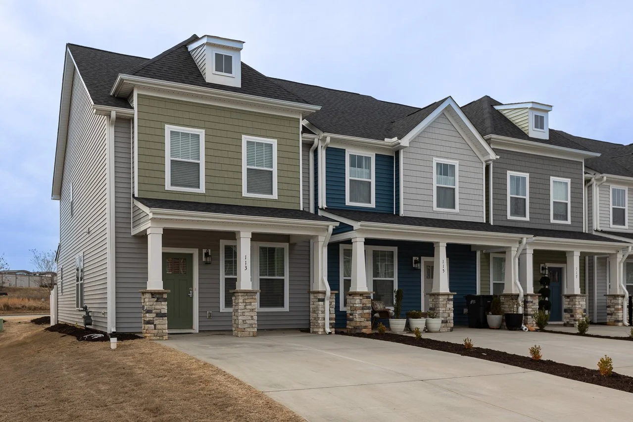 Front view of a modern townhouse complex with colorful exterior siding, stone accents on the porch columns, and a concrete driveway. The units have large windows and small front yards.