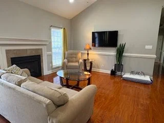 Living room with beige sofas, a wooden coffee table, a fireplace, a large flat-screen TV, a window with yellow curtains, a tall plant, and hardwood floors.
