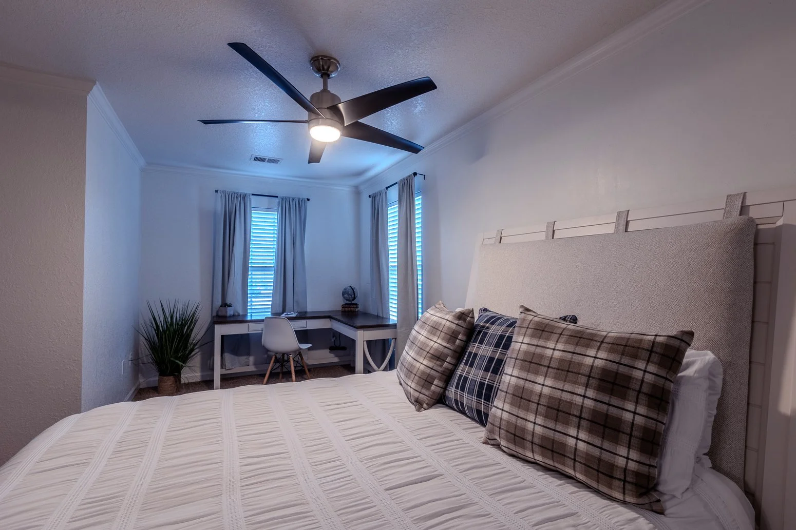 A bedroom with a white bed and plaid pillows, a ceiling fan, and a desk with a chair near windows with curtains.