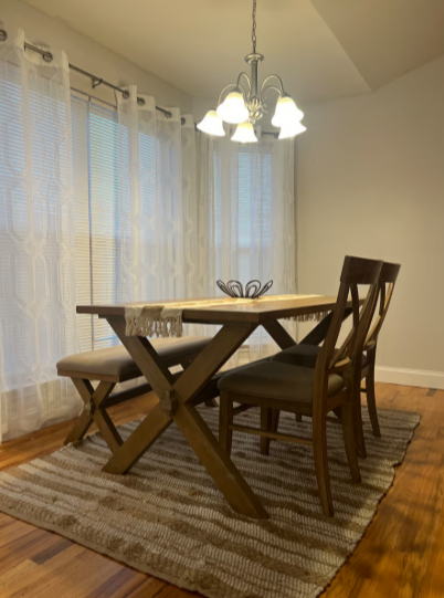 Dining room with wooden table, four chairs, chandelier, beige curtains, and a rug on hardwood floor.