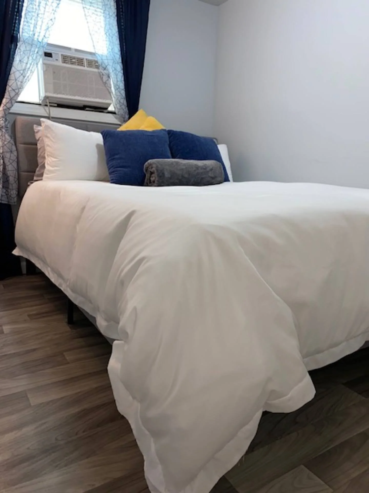 A neatly made bed with a white comforter, white pillows, dark blue pillows, yellow pillow, and a rolled gray blanket, in a room with wood flooring, blue curtains, and a window with an air conditioning unit.