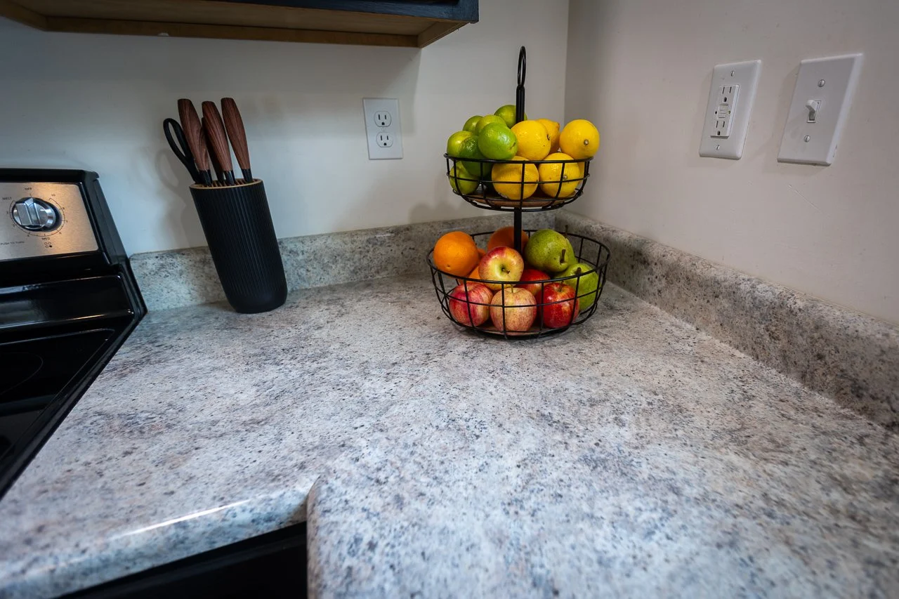 Countertop with a two-tier wire fruit basket holding apples, oranges, lemons, and limes, next to a knife holder and kitchen appliances.