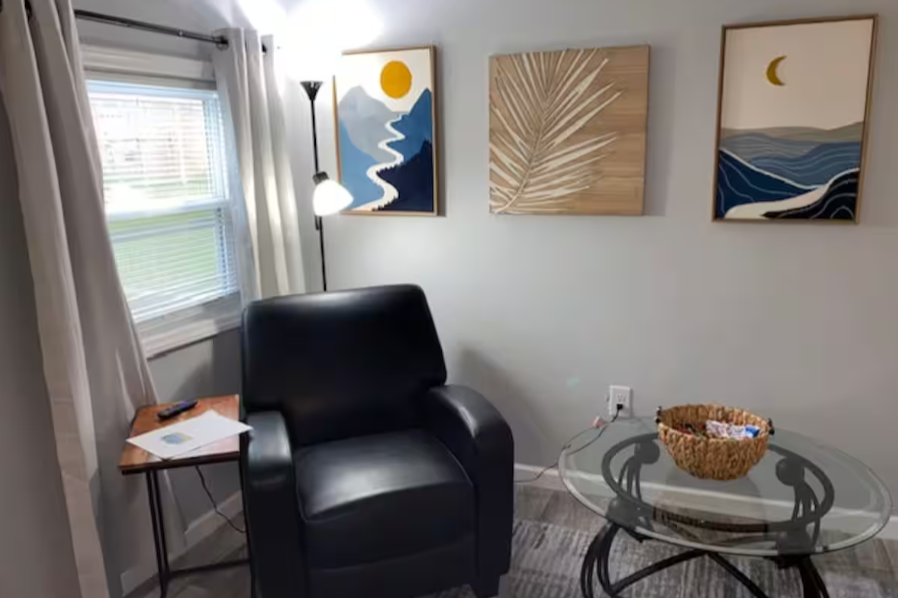 A cozy living room corner with a black leather armchair, a side table with papers and a remote, a glass coffee table with a basket, a standing lamp, a window with white curtains, and three abstract landscape pictures hanging on a white wall.